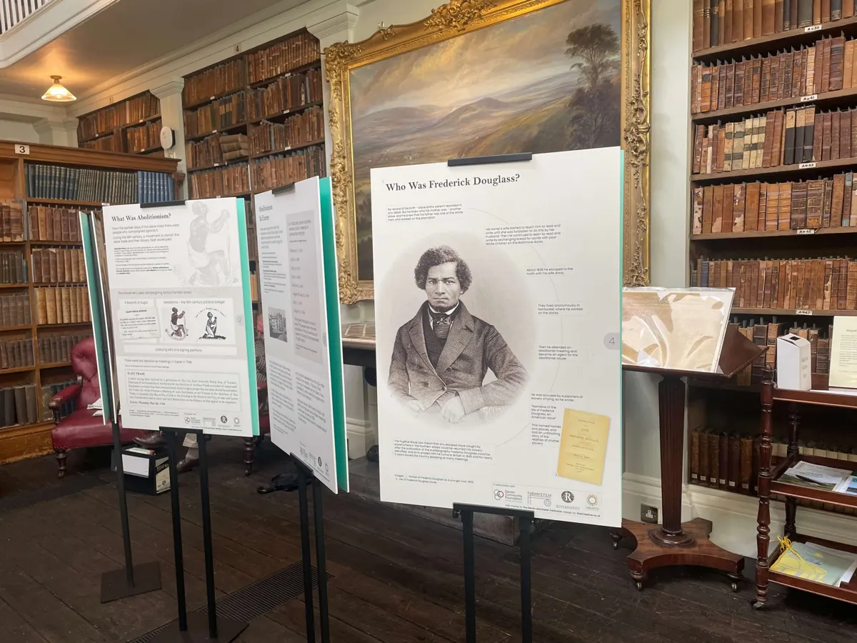 Display boards headed Who Was Frederick Douglass and What Was Abolitionism, standing in a library with tall wooden bookcases and an oil painting.